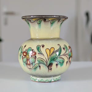 May include: A decorative, hand-painted ceramic vase with a cream-colored base and a flared rim. The vase features floral designs in shades of red, green, and yellow. The rim is trimmed with a brown and yellow pattern. The vase is approximately 7 inches tall.