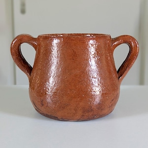 Può includere: Un vaso in ceramica marrone con due manici ad arco. Il vaso ha una superficie strutturata e una base arrotondata. L'interno è visibile e sembra essere dello stesso colore dell'esterno. Il vaso è appoggiato su una superficie bianca.