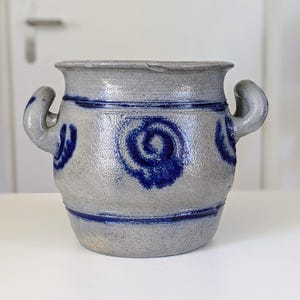 May include: A gray stoneware pot with two side handles, featuring a textured surface and cobalt blue painted spiral motifs around the body, accented by horizontal blue bands near the top and bottom rims of the vessel.