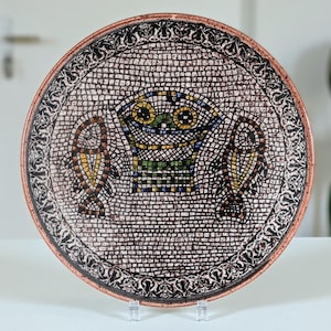Könnte beinhalten: Ein runder Keramikteller mit Mosaikdesign. Der Teller zeigt ein zentrales Bild eines Korbes und zweier Fische, alles im Mosaikstil. Der Rand hat ein dekoratives Muster. Der Teller wird auf einem transparenten Ständer ausgestellt.
