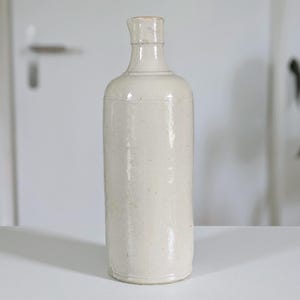 May include: A tall, cylindrical off-white ceramic bottle with a smooth, glossy finish. The bottle features a narrow neck, a slightly flared rim, and a flat base. The surface has subtle variations in color and texture, indicating a handcrafted appearance.