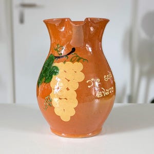 Puede incluir: Una jarra de cerámica con un esmalte naranja cálido. La jarra está decorada con un racimo de uvas amarillas y hojas verdes. El texto "De Sois" y "Mare" está escrito en la jarra.
