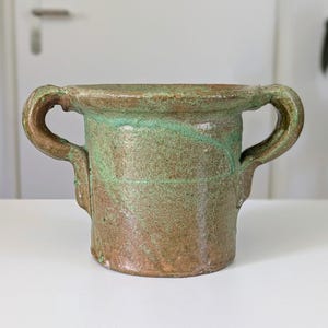 May include: A ceramic vessel with two side handles, featuring a textured surface with mottled brown and green glaze. The pot has a wide, flared rim and a cylindrical body, showcasing a weathered appearance with visible variations in colour and finish.
