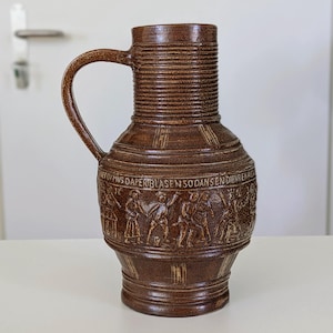 以下が含まれることがあります： リブ付きの首とハンドルが付いた茶色の陶器製ピッチャー。胴体には動きのある人物を描いたレリーフ帯があり、「DIEV MUS DAPER BLASEN SO DANS EN DIEVRENAST」という文字が刻まれています。表面は斑点のある釉薬仕上げです。