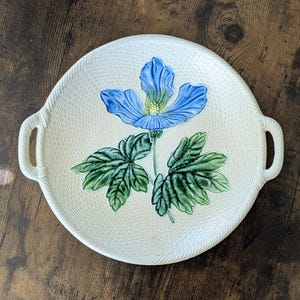 May include: A round, cream-colored ceramic serving tray with two side handles. The surface features a textured, woven-like pattern and a central hand-painted illustration of a large blue flower with a yellow center and green leaves.