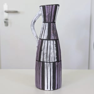Könnte beinhalten: Ein hoher Keramikkrug mit einer dunkel schwarzen Grundfarbe und vertikalen Streifen in hellen Violett- und Weißtönen. Der Krug verfügt über einen einzelnen geschwungenen Henkel an der Seite und einen ausgestellten Rand mit strukturierter Oberfläche.