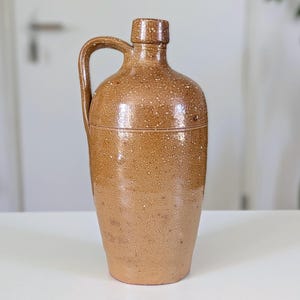 Puede incluir: Una jarra de gres alta y cilíndrica con un asa lateral y un cuello estrecho. El exterior presenta un esmalte moteado en tonos beige y marrón claro, con una línea horizontal que rodea la parte superior del cuerpo, dándole un aspecto terroso.