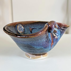 May include: A handmade ceramic bowl with a unique handle. The bowl features a blend of blue, purple, and brown hues with a glossy finish. The rim and handle have a darker brown color, and the base is a light beige.
