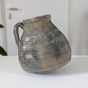 May include: A rustic, gray-toned ceramic jug with a handle. The jug has a wide body and a narrow neck, with visible horizontal ridges. The surface has a weathered appearance, suggesting age and use. The jug is likely for decorative or functional purposes.