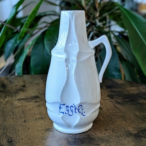 May include: A white ceramic pitcher featuring a glossy finish and a distinct molded geometric pattern on its body. The word Effie is written in blue cursive script on the front. It includes a single handle on the right side and a flared rim.