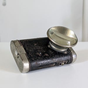 May include: A vintage rectangular flashlight with a black textured body and silver metal trim. It features a prominent, circular, convex glass lens mounted on the top side, along with a small metal switch on the side of the casing.