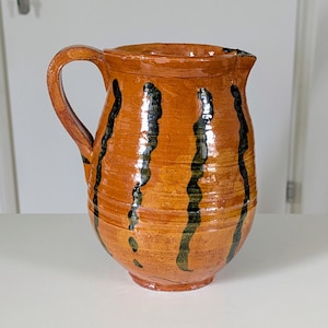 May include: A handmade ceramic pitcher with a wide base and a curved handle. The pitcher is glazed in a warm orange color with vertical black stripes. It has a glossy finish and a spout for pouring.