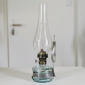 Puede incluir: Una lámpara de aceite de vidrio transparente con un marco de metal plateado y un asa redonda. La lámpara tiene una base bulbosa y una chimenea alta y delgada. La lámpara está sobre una superficie blanca.