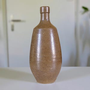Puede incluir: Una botella alta de gres marrón con cuello estrecho y cuerpo redondeado. La botella tiene una superficie texturizada y líneas horizontales cerca del cuello. Se muestra sobre una superficie blanca.