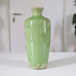 Puede incluir: Un jarrón cilíndrico alto de cerámica con un esmalte verde claro brillante que presenta un patrón de grietas oscuras en toda la superficie. El jarrón tiene un cuello estrecho, un borde acampanado y una base texturizada y desgastada.