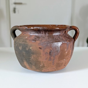 Könnte beinhalten: Ein rustikaler, brauner Keramiktopf mit zwei Henkeln. Der Topf hat einen abgerundeten Körper und eine weite Öffnung. Die Außenseite weist ein gesprenkeltes, gealtertes Aussehen mit Farbvariationen auf, darunter Brauntöne und rötlich-braune Farbtöne. Der Topf ist wahrscheinlich Vintage.