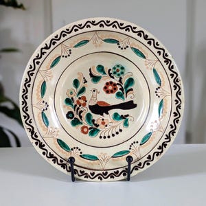 Puede incluir: Un plato de cerámica color crema con un diseño pintado a mano. El plato presenta una imagen central de un pájaro rodeado de flores en tonos marrones, verdes y naranjas. El borde está decorado con un patrón repetitivo de remolinos negros y crema y hojas verdes.