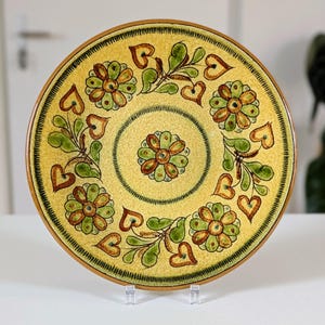 May include: A decorative ceramic plate with a yellow background and a floral design. The plate features brown, green, and yellow flowers, green leaves, and brown heart shapes. The rim is brown, and the plate is on a clear stand.