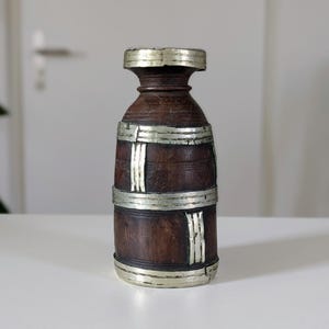 May include: A dark brown wooden vase with silver metal bands and accents. The vase has a cylindrical shape with a slightly wider base and top. The silver bands are placed around the middle and the top and bottom of the vase.