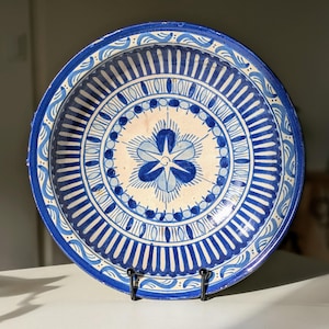 Puede incluir: Un plato redondo de cerámica pintado a mano con un diseño floral azul y blanco. El plato presenta un motivo floral central rodeado de círculos concéntricos y líneas radiales. El borde tiene un patrón ondulado.