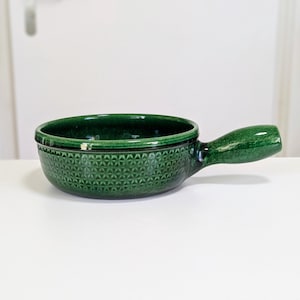 May include: A dark green ceramic bowl with a handle. The bowl has a textured pattern and a glossy finish. The handle is rounded and attached to the side of the bowl. The bowl is sitting on a white surface.