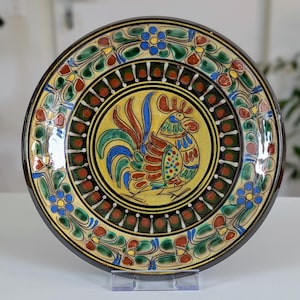 May include: A decorative ceramic plate with a hand-painted rooster design. The plate features a yellow center with a rooster, surrounded by a black border and colorful floral patterns in green, red, and blue. The plate has a dark brown rim.