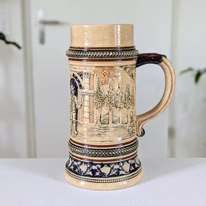 Può includere: Un boccale di birra alto in ceramica beige con manico. Il boccale presenta dettagliate opere d'arte in rilievo raffiguranti una scena con figure, alberi ed elementi architettonici. Ha fasce decorative marroni e blu e un coperchio. Il manico è marrone scuro.