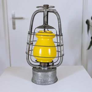 May include: A vintage-style metal lantern featuring a weathered grey finish and a bright yellow glass globe. The lantern is encased in a protective wire cage with a handle at the top and a circular base, showcasing a classic design with visible signs of age.