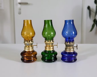 Puede incluir: Tres lámparas de aceite pequeñas con depósitos y chimeneas de vidrio en color ámbar, verde y azul. Cada lámpara cuenta con un quemador de metal color latón con una pequeña perilla de ajuste en el lateral, uniendo la base con la chimenea.