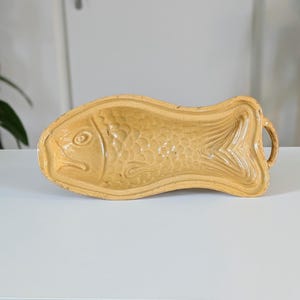 以下が含まれることがあります： 詳細な鱗とひれが付いた、魚の形をした薄茶色のセラミック型。型の両側に取っ手があり、焼くか提供するように設計されています。セラミックの作品は白い表面に置かれています。