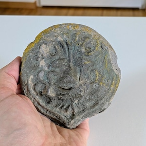 May include: A weathered, round, gray stone carving of a face with detailed features. The stone has areas of yellow and green discoloration, suggesting age and exposure to the elements. The carving is held in a hand.