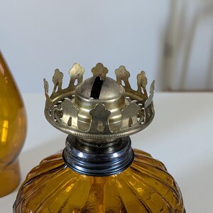 May include: Close-up of a vintage amber glass oil lamp with a decorative brass crown-shaped top. The lamp has a ribbed, textured glass base and a metal wick holder. A matching amber glass vase is partially visible in the background.