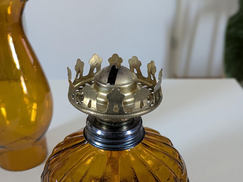 May include: Close-up of a vintage amber glass oil lamp with a decorative brass crown-shaped top. The lamp has a ribbed, textured glass base and a metal wick holder. A matching amber glass vase is partially visible in the background.