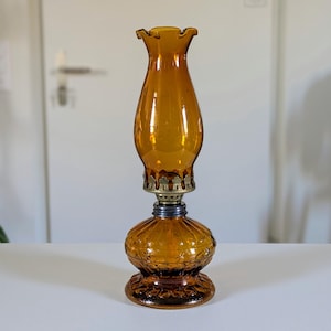 May include: Amber-colored glass oil lamp with a ruffled top and ornate base. The lamp features a brass-colored metal fitting and a dark blue ring. The lamp is a vintage style and sits on a white surface.