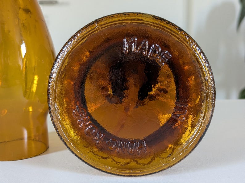 May include: Close-up of the bottom of a brown glass bottle. The base is circular with the embossed text "MADE IN HONG KONG" in a circular pattern. The bottle has a glossy, amber color.