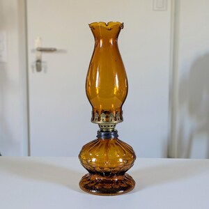 May include: Amber-colored glass oil lamp with a ruffled top and ornate base. The lamp features a brass collar and a dark blue accent ring. The glass has a textured, ribbed design. A classic decorative piece.