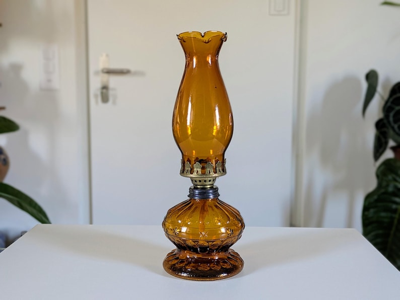 May include: Amber-colored glass oil lamp with a ruffled top and ornate base. The lamp features a brass collar and a dark blue accent ring. The glass has a textured, ribbed design. A classic decorative piece.