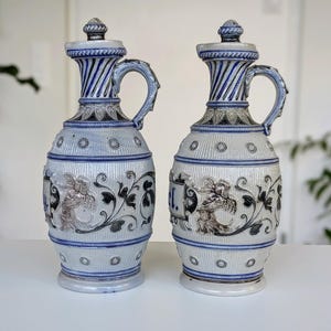 Puede incluir: Dos jarras antiguas de cerámica con tapas. Las jarras son blancas con detalles azules, incluyendo rayas en el cuello y el asa. Cada jarra presenta un diseño en relieve detallado de figuras y follaje. Las jarras se exhiben sobre una superficie blanca.