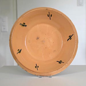 May include: A round, light orange ceramic bowl with a smooth interior surface. The rim features a series of small, evenly spaced black painted markings that resemble stylized dashes or abstract symbols arranged in a circular pattern.