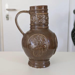 May include: A brown glazed ceramic jug featuring a rounded body with an embossed coat of arms and the text VANEYEN WIEGPRECHT. The neck is decorated with a relief pattern of leaves and flowers, and it includes a single curved handle.