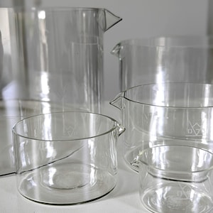 Puede incluir: Una colección de vasos de precipitados de vidrio transparente de varios tamaños. Cada vaso tiene un pico y una base plana. Los vasos están dispuestos en grupo, algunos se superponen. El vidrio transparente permite ver a través de los recipientes. Los vasos son probablemente para uso en laboratorio o cocina.