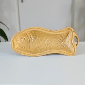 Peut inclure: Un moule en céramique en forme de poisson de couleur beige, avec des écailles et des nageoires détaillées. Le moule a une poignée de chaque côté et est conçu pour la cuisson ou le service. La pièce en céramique est posée sur une surface blanche.
