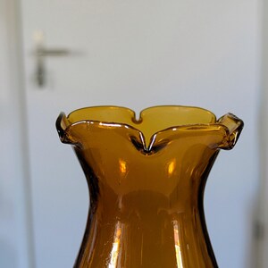 May include: Amber-colored glass vase with a ruffled rim. The vase has a flared shape and a smooth, glossy surface. The rim has a scalloped edge, adding a decorative touch. The vase is likely vintage or retro in style.