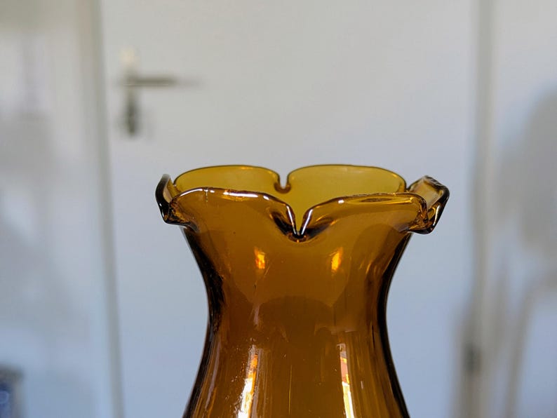 May include: Amber-colored glass vase with a ruffled rim. The vase has a flared shape and a smooth, glossy surface. The rim has a scalloped edge, adding a decorative touch. The vase is likely vintage or retro in style.