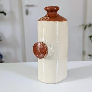 Peut inclure: Une bouteille en céramique blanc cassé avec un dessus marron et un élément circulaire marron décoratif sur le côté. La bouteille a une forme cylindrique avec une surface légèrement texturée. Design simple et rustique, pour la décoration intérieure.