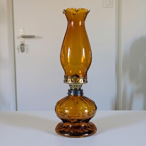 May include: Amber-colored glass oil lamp with a ruffled top and ornate base. The lamp features a brass-colored metal accent and a blue-colored metal connector. The lamp is a vintage style and sits on a white surface.