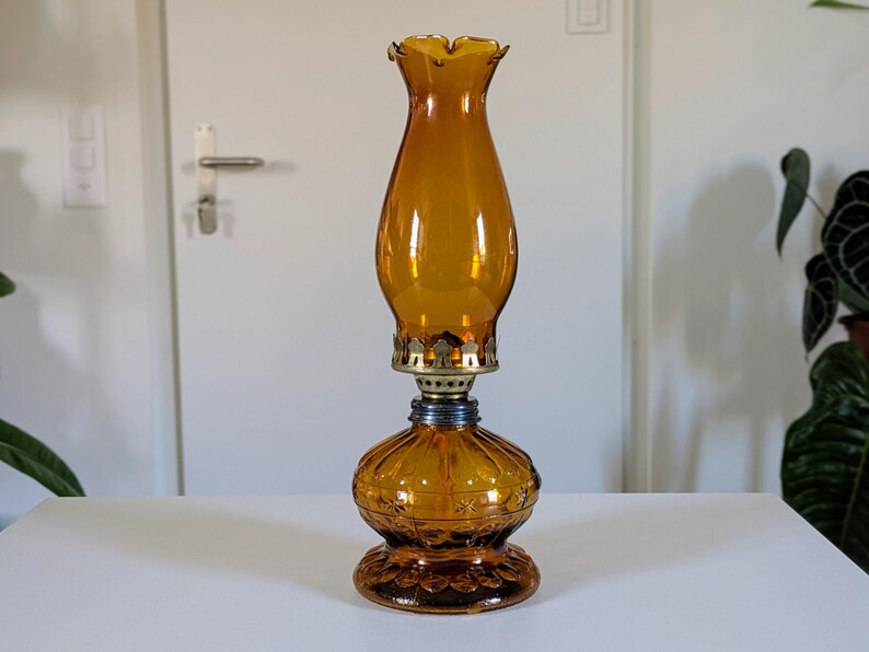 May include: Amber-colored glass oil lamp with a ruffled top and ornate base. The lamp features a brass-colored metal accent and a blue-colored metal connector. The lamp is a vintage style and sits on a white surface.