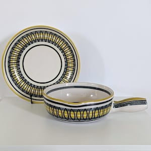 May include: A ceramic bowl with a handle and a matching plate. Both items feature a white base with a yellow, black, and grey geometric design. The plate is round, and the bowl is oval with a handle. The set is on a white surface.