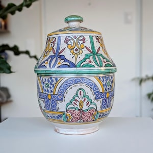 Może przedstawiać: Ceramiczny słoik z pokrywką, z kolorowym, skomplikowanym wzorem. Słoik jest głównie biały z niebieskimi, żółtymi, zielonymi i czerwonymi wzorami. Pokrywka ma mały uchwyt. Słoik stoi na białej powierzchni.