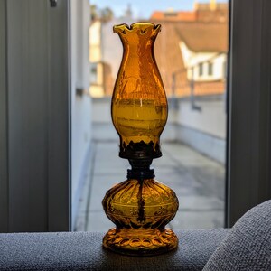 May include: An amber-colored glass oil lamp with a ruffled top and ornate base. The lamp has a dark metal connector and a decorative floral pattern on the base. The lamp is sitting on a gray surface in front of a window.